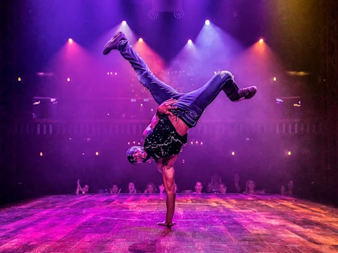A dancer performs a one-handed handstand on stage under colorful spotlights, with an audience watching in the background.