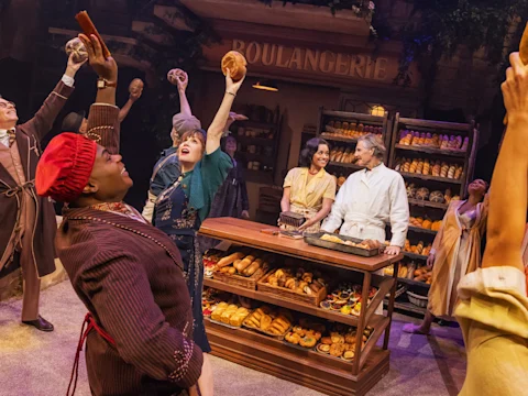 Production photo of The Baker's Wife in New York, showing actors on stage perform in a bakery set, holding up bread and pastries, with shelves of baked goods and a "BOULANGERIE" sign in the background.