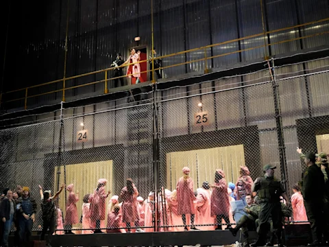 A group of people in pink robes stand behind a metal fence while a figure in red stands above on a platform, with uniformed guards at the front.