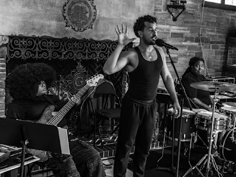 A man sings into a microphone with one hand raised, accompanied by a bassist and a drummer, performing indoors in front of a fireplace.