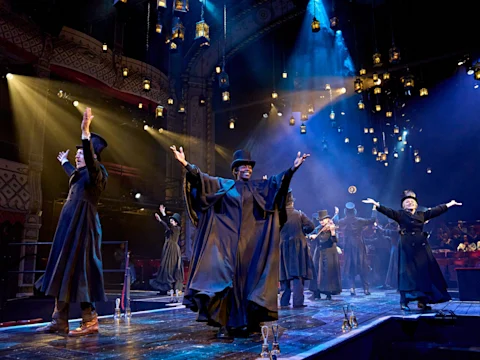 Actors in dark costumes and top hats perform on a theater stage, arms raised, with hanging lanterns and a dramatic, ornate backdrop.
