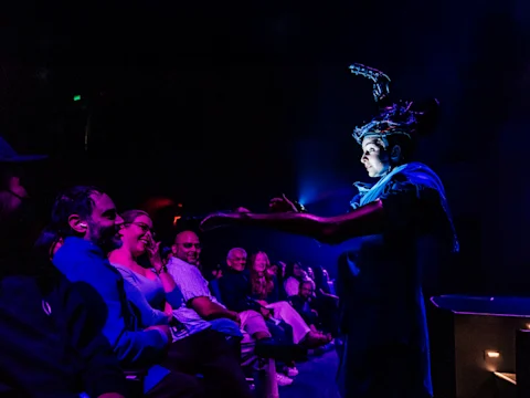 Production photo of ha ha ha ha ha ha ha in Pasadena, showing a performer in a costume interacts with an audience seated in a dimly lit theater, with blue and purple stage lighting.