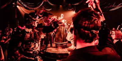 People in ornate masquerade costumes and masks dance at a dimly lit, dramatic event; a person stands spotlighted on a central platform.