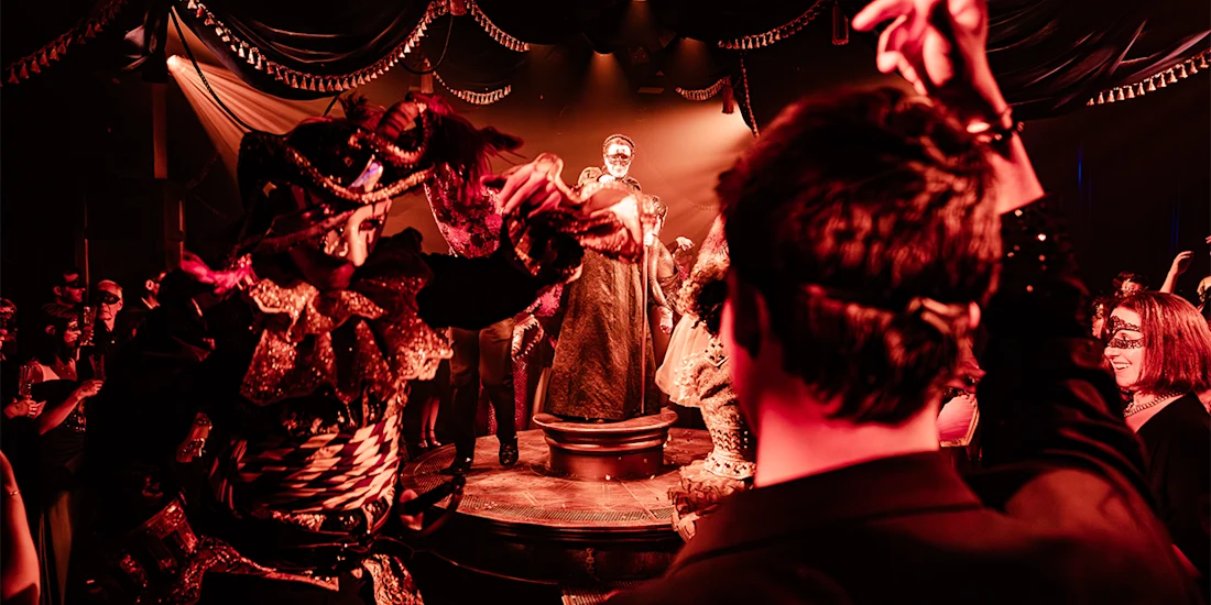 People in ornate masquerade costumes and masks dance at a dimly lit, dramatic event; a person stands spotlighted on a central platform.