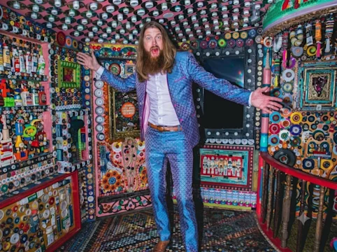 A man in a patterned blue suit stands with arms outstretched in a colorful, art-filled room decorated with buttons, bottle caps, and other objects on the walls, floor, and ceiling.