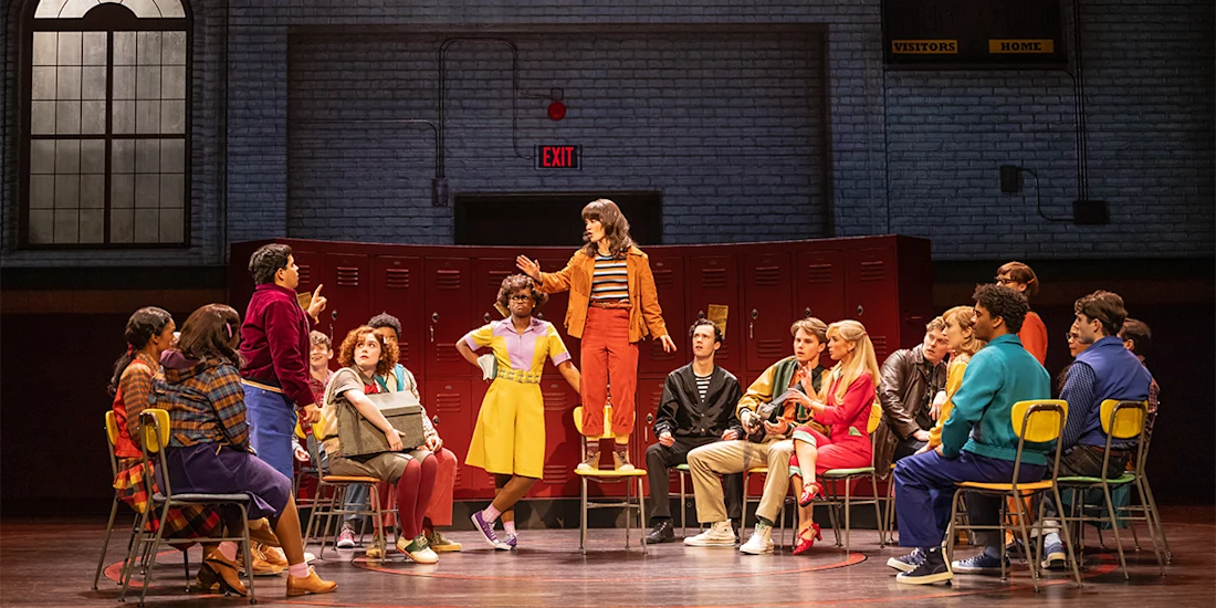 A group of actors in colorful 1980s-style clothing sit on chairs in a semicircle on stage, with one person standing on a chair speaking, set against red lockers and a gymnasium background.
