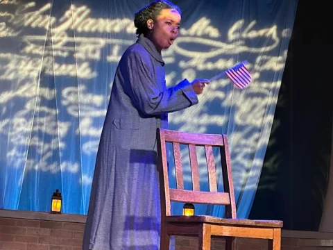 A person in a blue coat gestures while holding a small American flag. They stand next to a wooden chair, with handwritten cursive text projected on a screen behind them.