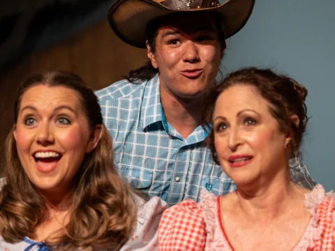 Three people are posing together, two women in front and one man in a cowboy hat behind them. All three have cheerful or playful expressions.