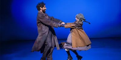 Two actors in period costumes hold hands and spin each other on stage against a blue background.