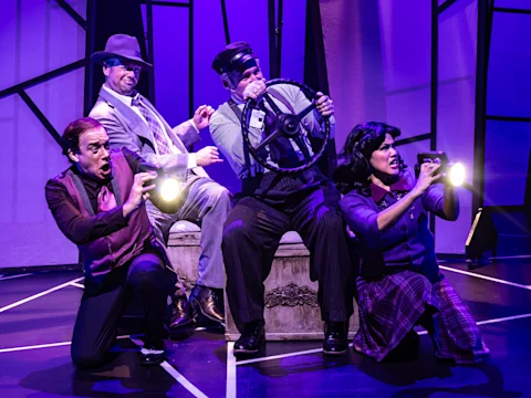 Four actors perform on stage in dramatic lighting, miming a car ride with props and expressive poses against a geometric purple background.
