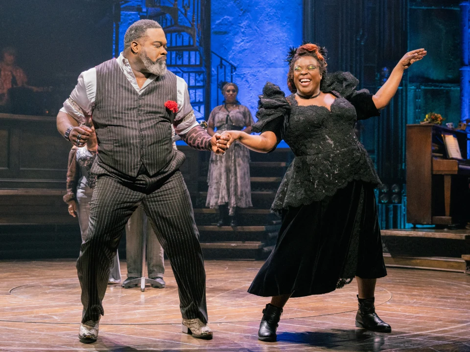 Two actors perform a lively dance on stage in period costumes, with the man in a pinstripe suit and the woman in a black dress, set against a vintage backdrop with dim lighting.