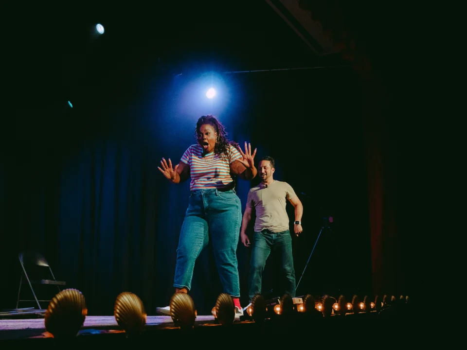 Two people perform on stage with dramatic expressions. One person stands in the foreground with hands up, and the other is slightly behind. Stage lighting illuminates the scene.