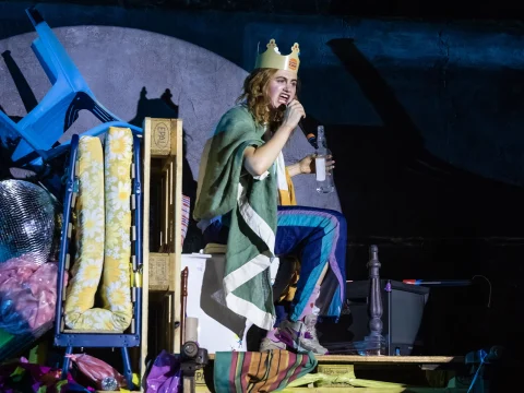 Person wearing a paper crown sits among cluttered objects, holding a bottle and wearing a green-striped blanket.