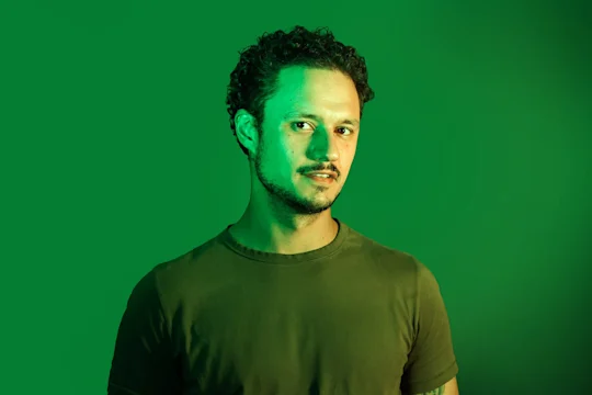 A man with short curly hair and a trimmed beard stands in front of a green background, wearing a green t-shirt, looking slightly to the side under green lighting.