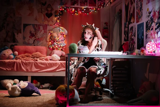 Person in a cow-patterned outfit sits at a desk with a headset in a room decorated with anime posters, plush toys, and string lights.