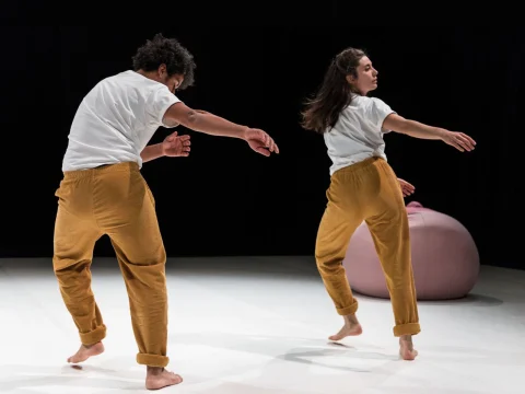 Two dancers in white shirts and mustard pants perform on stage with a large pink object in the background.