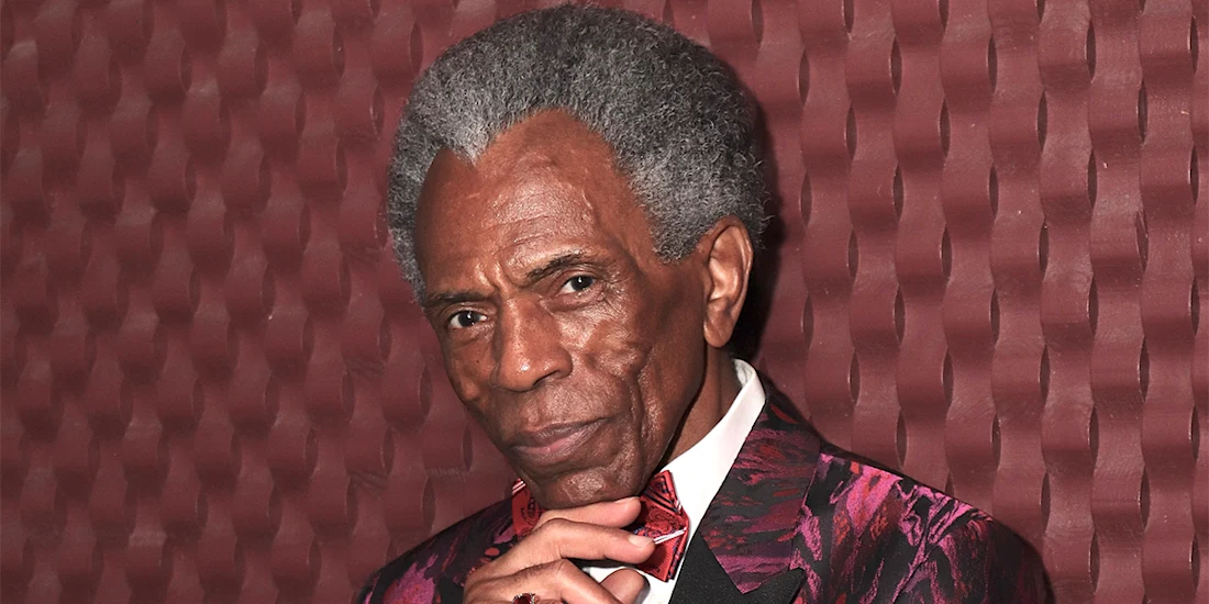 An older man with gray hair wearing a patterned burgundy suit and bow tie stands against a textured maroon wall, resting his chin on his hand.