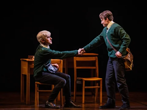 Production photo of Harry Potter and the Cursed Child in Costa Mesa showing two people in green sweaters shake hands; one is seated at a desk while the other stands with a satchel over his shoulder.