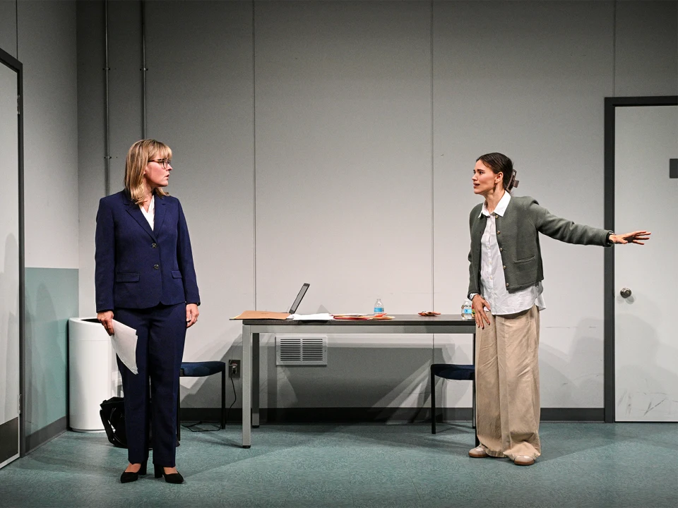 Two women stand in an office; one in a suit holds papers, the other in casual attire gestures. A desk with a laptop and water bottle is in the background.