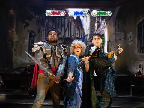 Three actors in medieval costumes on stage, portraying characters with weapons in a dramatic setting, under theatrical lighting.