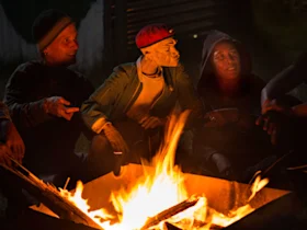 A group of people and a puppet sit around a bonfire at night.