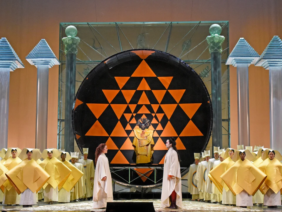 Stage scene with performers in robes, a large black circular structure with orange geometric patterns, and group of people in yellow costumes. Four tall columns with decorative tops frame the stage.