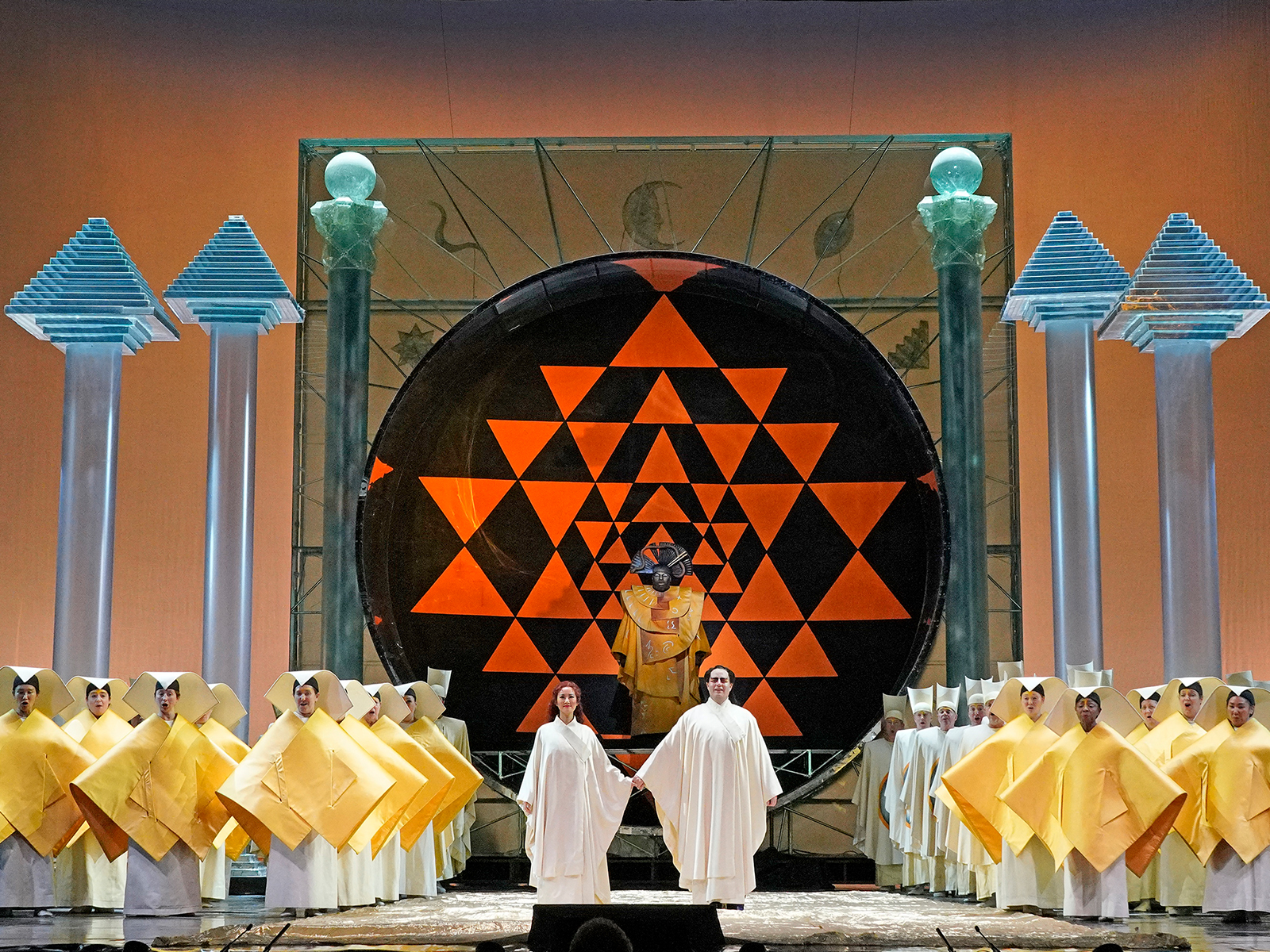 Stage scene with performers in robes, a large black circular structure with orange geometric patterns, and group of people in yellow costumes. Four tall columns with decorative tops frame the stage.