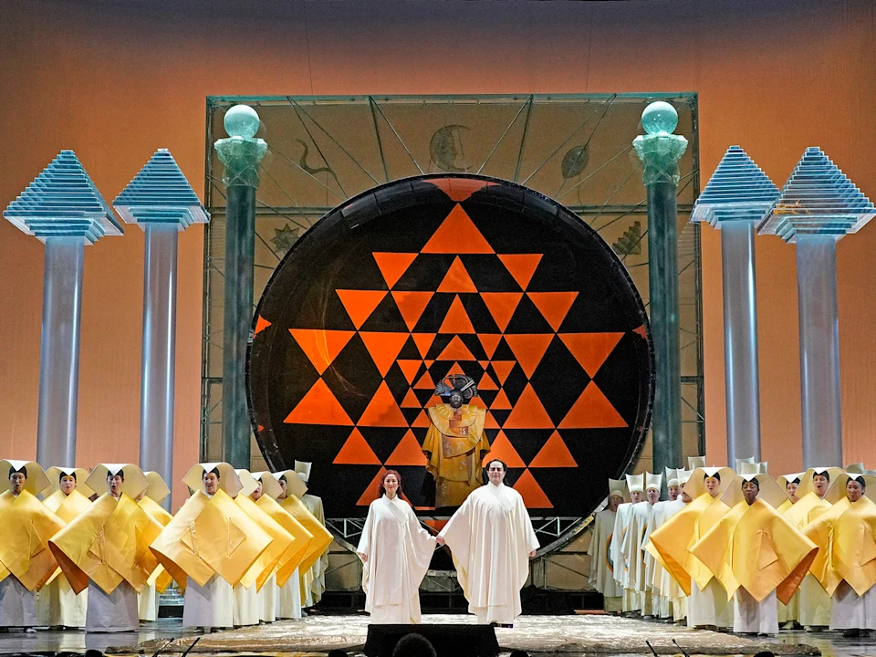 Stage scene with performers in robes, a large black circular structure with orange geometric patterns, and group of people in yellow costumes. Four tall columns with decorative tops frame the stage.