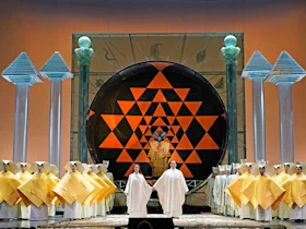 Stage scene with performers in robes, a large black circular structure with orange geometric patterns, and group of people in yellow costumes. Four tall columns with decorative tops frame the stage.