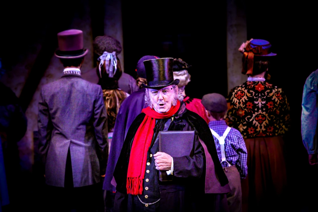 An actor in a theatrical costume, wearing a top hat and red scarf, holds a book while performing on stage with a group of people in period attire.