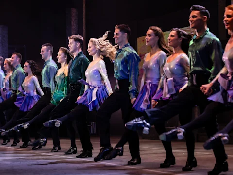 A group of dancers in coordinated costumes performs a high-kick routine on stage, with synchronized movements and stage lighting.