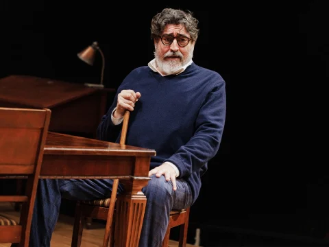 An older man with glasses and a beard, wearing a navy blue sweater, sits at a wooden desk holding a cane. A small lamp is seen on the desk behind him.