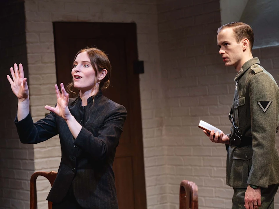 Two actors perform in a scene; a woman gestures expressively while a man in a military uniform watches, holding a book.