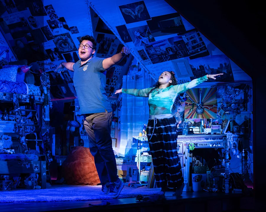 Two people stand with arms outstretched in a blue-lit, poster-covered room, appearing to perform on stage amid cluttered furniture and decorations.