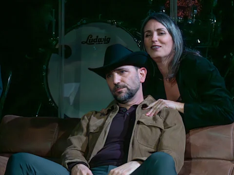 A man in a cowboy hat sits on a couch looking serious, while a woman stands behind him with her hands on his shoulders, both in front of a drum set.