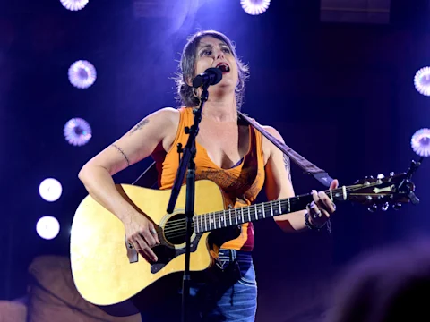 A woman in an orange tank top plays an acoustic guitar and sings into a microphone on stage, with stage lights in the background.