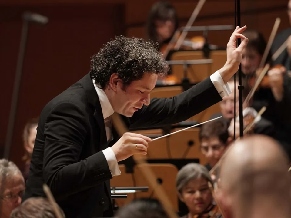 Conductor leading an orchestra, holding a baton and gesturing expressively, with musicians playing string instruments in the background.