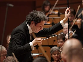Conductor leading an orchestra, holding a baton and gesturing expressively, with musicians playing string instruments in the background.