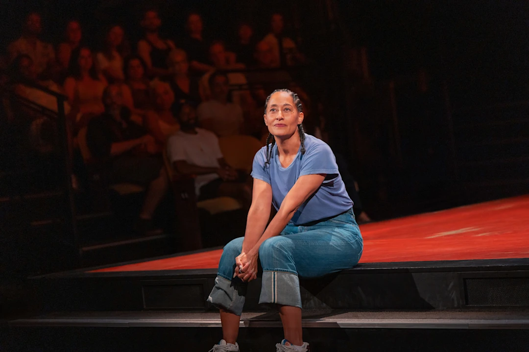 A woman in a blue shirt and rolled-up jeans sits on stage with her hands clasped on her knees, facing the audience seated in the background.