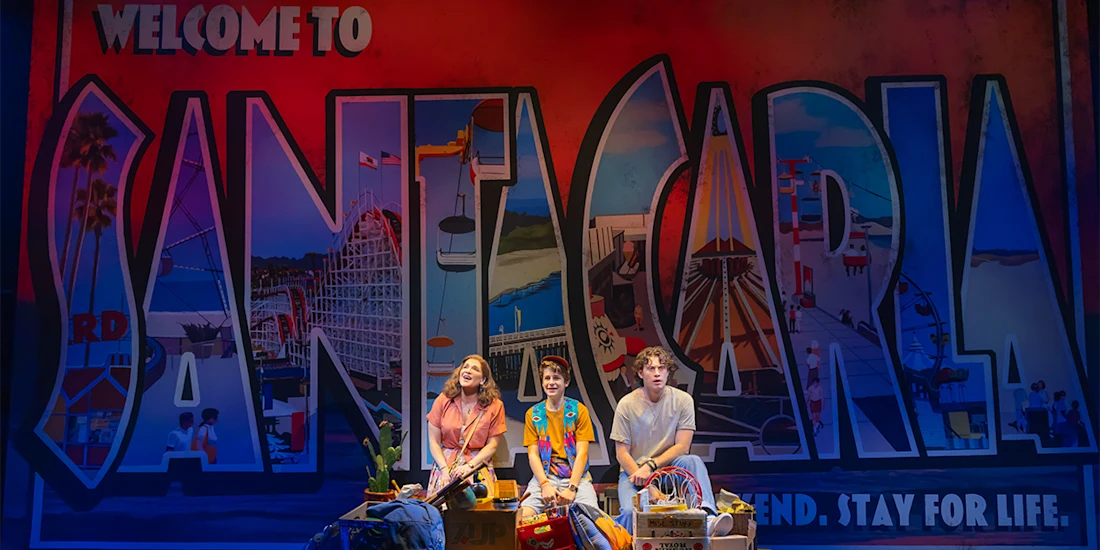 Three people sit on a stage set in front of a large "Welcome to Santa Carla" mural, with colorful graphics and beach-themed props around them.