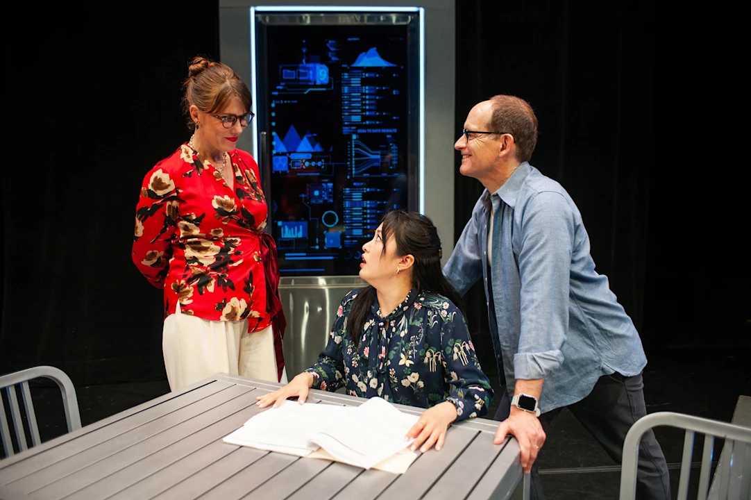 Three adults converse around a table with papers, while a digital screen displaying data is visible in the background.