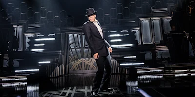 A person in a suit and fedora performs a dance move on a dimly lit stage with art deco-style set designs and spotlights.