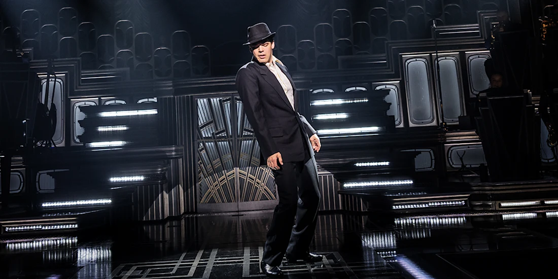 A person in a suit and fedora performs a dance move on a dimly lit stage with art deco-style set designs and spotlights.