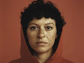 A person with short curly hair and freckles wears a red hoodie and looks directly at the camera, set against a plain red background.