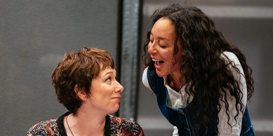 Two women interact; one with short hair sits and looks up while the other, with long curly hair, leans over and speaks animatedly to her.