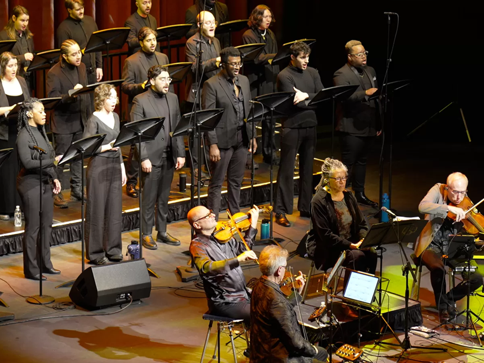 A choir dressed in black performs on stage with orchestral musicians seated in front playing string instruments.