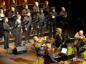 A choir dressed in black performs on stage with orchestral musicians seated in front playing string instruments.