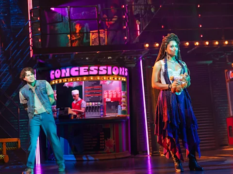 Two actors perform on stage in front of a brightly lit concessions stand with snacks and a menu, while another person works behind the counter.