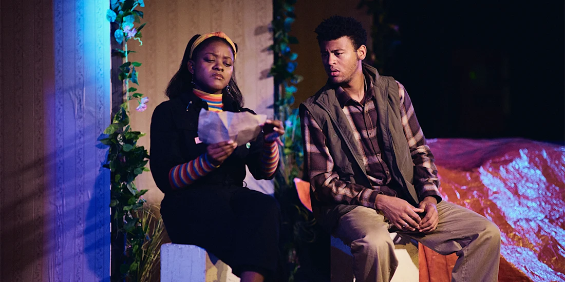 Two people sit next to each other; one reads a sheet of paper intently while the other looks on, both surrounded by foliage and stage props.