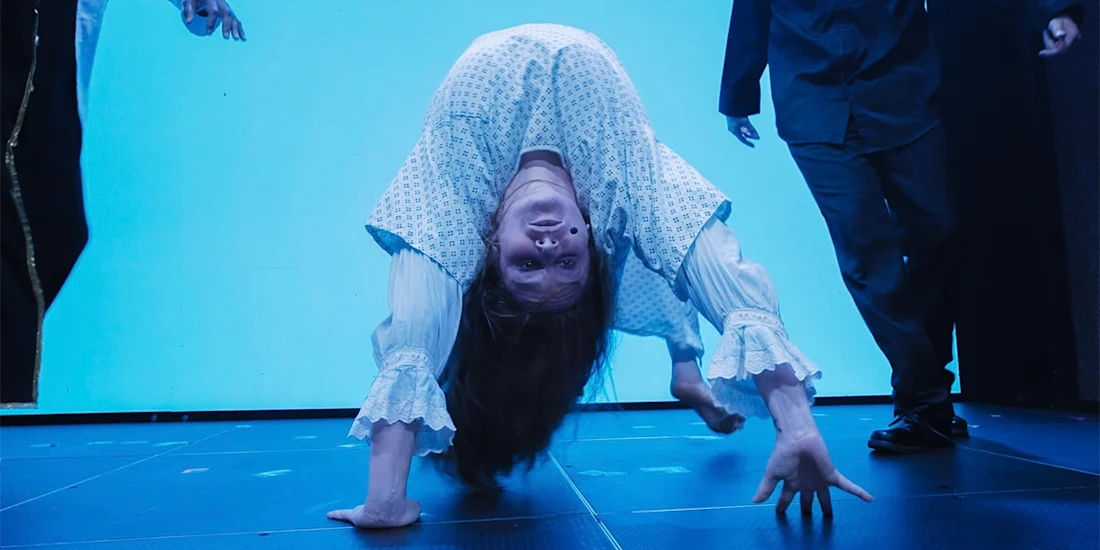 A person in a white gown bends backward in an unnatural pose on stage, with hair hanging down and hands touching the floor. Two other people stand nearby.